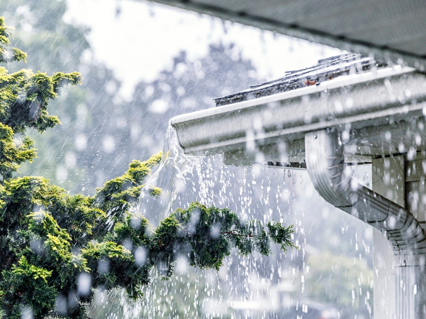 Drenching downpour rain storm water is overflowing off the tile shingle roof - streaming, rushing and splashing out over the overhanging eaves trough aluminum roof gutter system on a suburban residential colonial style house near Rochester, New York State, USA during a torrential mid-summer July downpour.