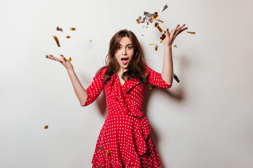 Joyful brunette in red dress scatters gold and silver confetti on white background