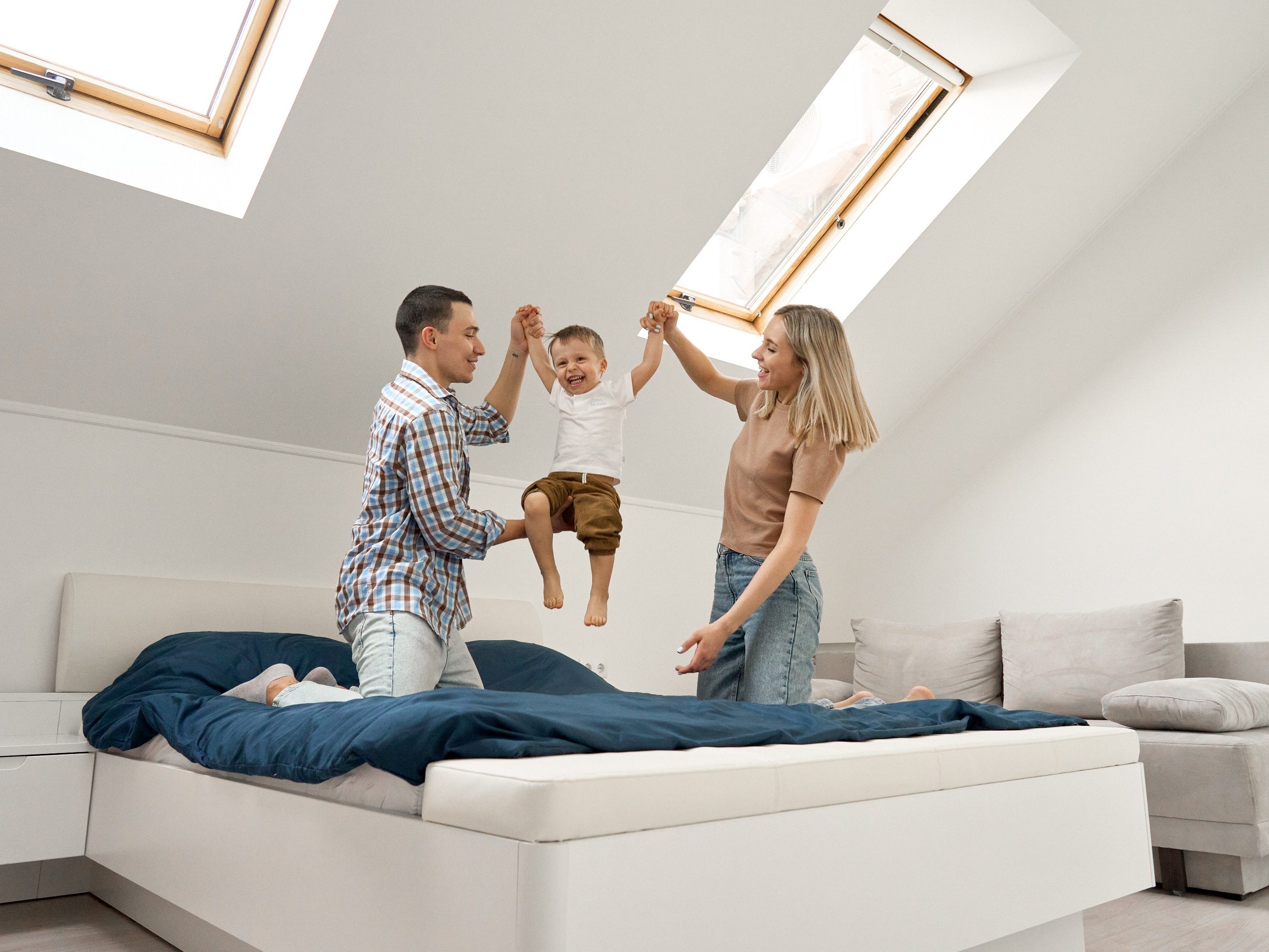 Happy young family couple playing with cute small toddler kid son in bedroom at home. Parents and child boy having fun feeling joy jumping on bed bonding spending weekend morning time together.