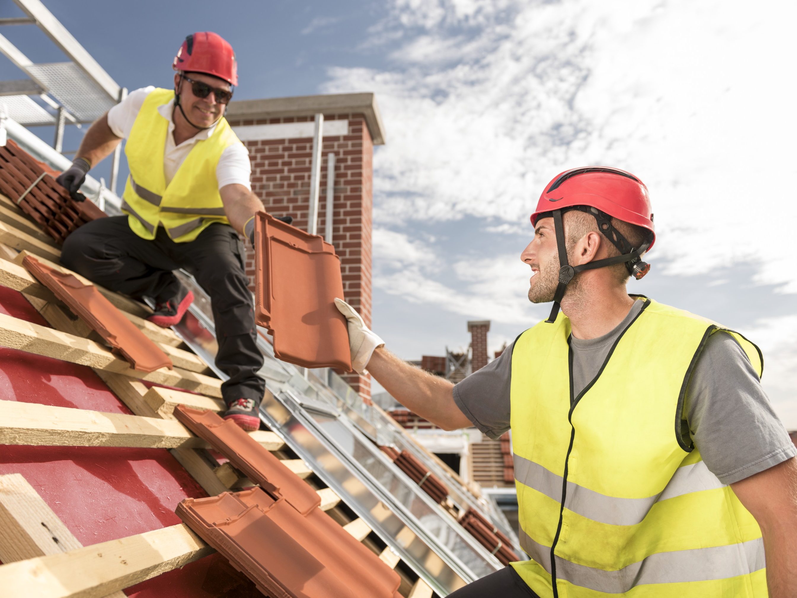 Urban roofers passing on roof tile towards colleague working on the roof wearing safety jackets and hard hats