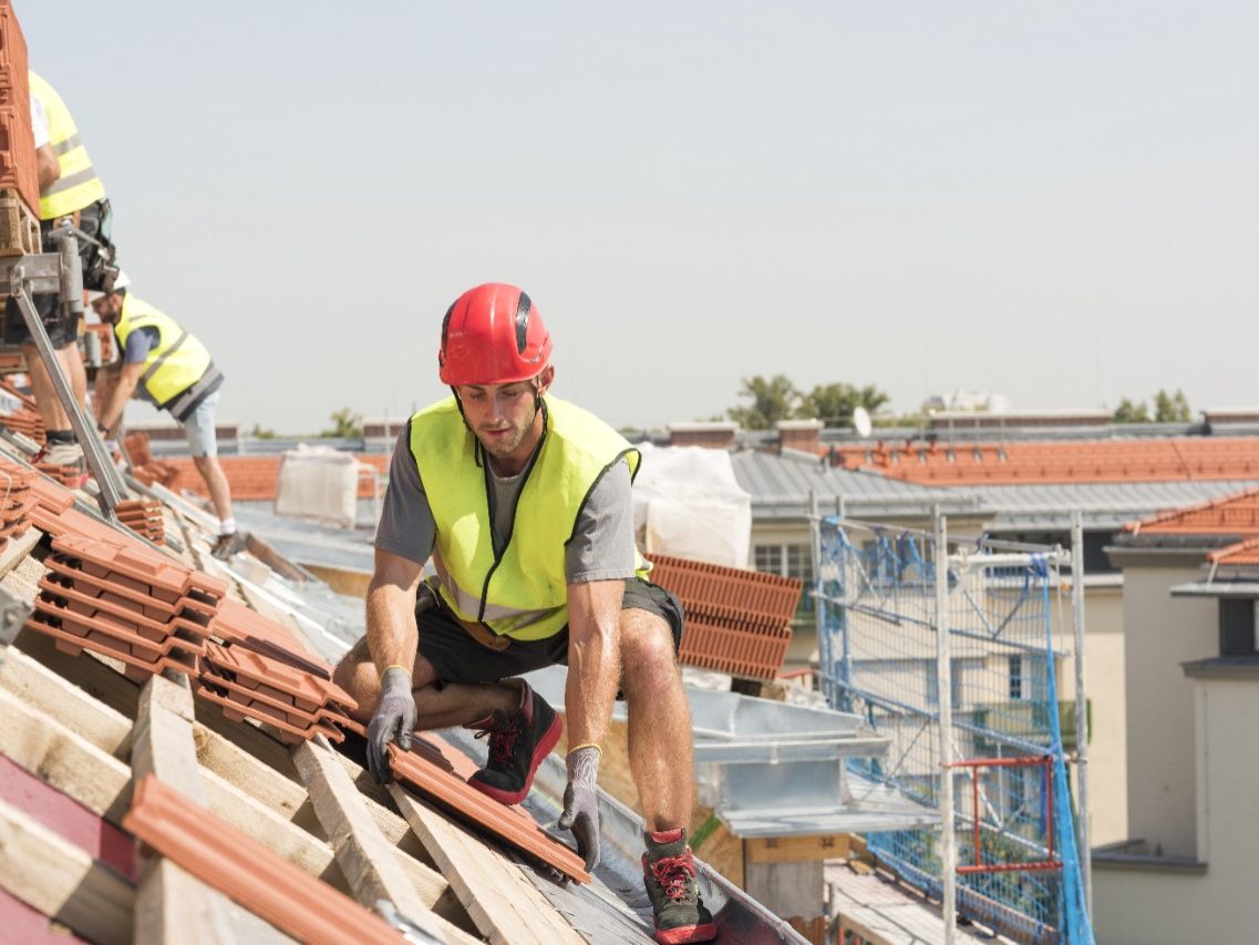 Urban roofers at work wearing safety jacket and hard hat laying roof tiles at an urban location