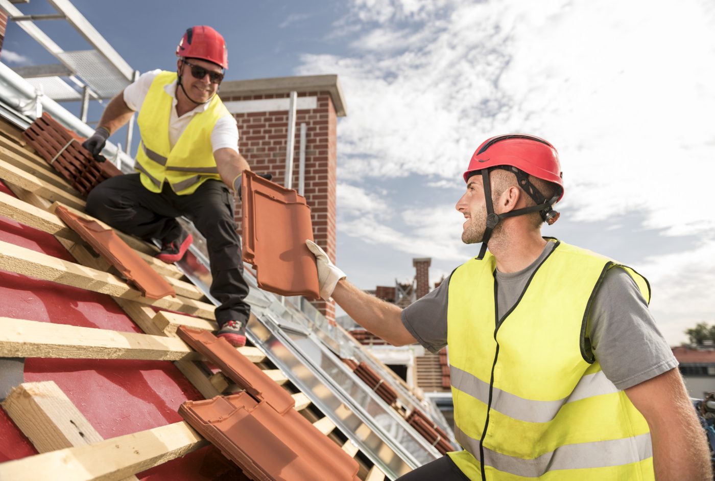 Urban roofers passing on roof tile towards colleague working on the roof wearing safety jackets and hard hats