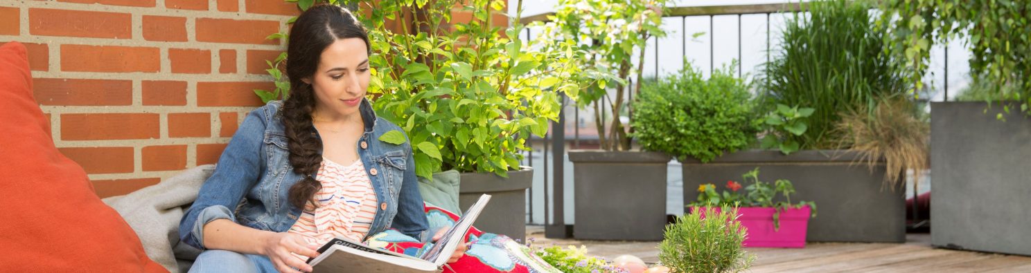 Woman surrounded by colorful cushions and blankets on rooftop terrace flipping through photo album in front of brick wall